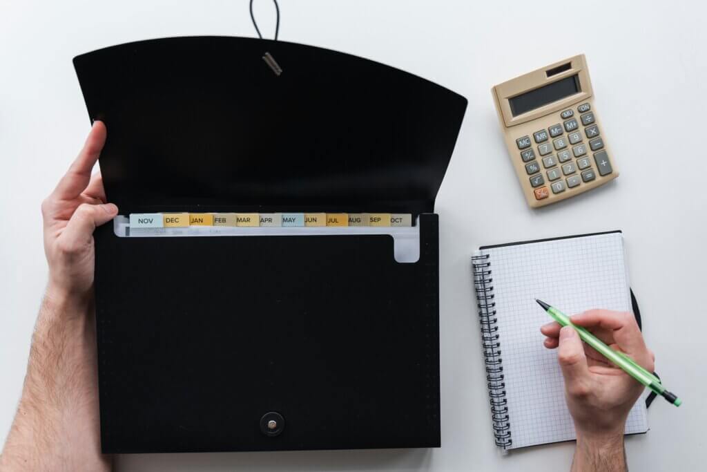 Hands holding open a black file folder with month dividers. A notebook, pen, and calculator are nearby on a white surface.