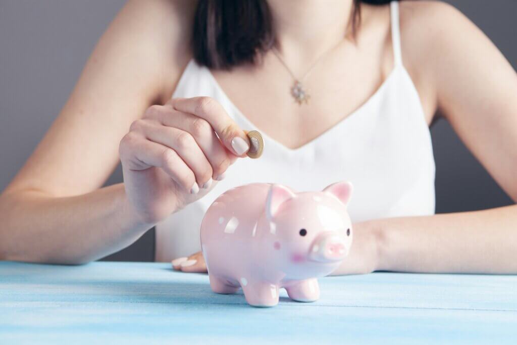 Closeup of woman in a white camisole putting a coin into a pink piggy bank
