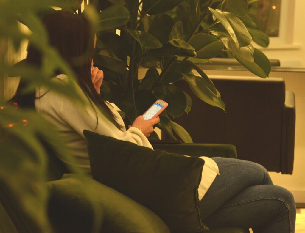 Girl using smartphone sitting on a black sofa surrounded by green plants.